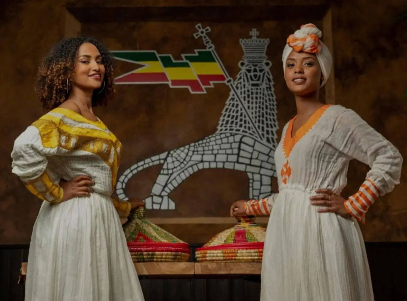 Zwei Frauen im traditionellen Habesha-Kemis vor dem Wandbild mit der Stele von Aksum, davor zwei Mesob-Körbe