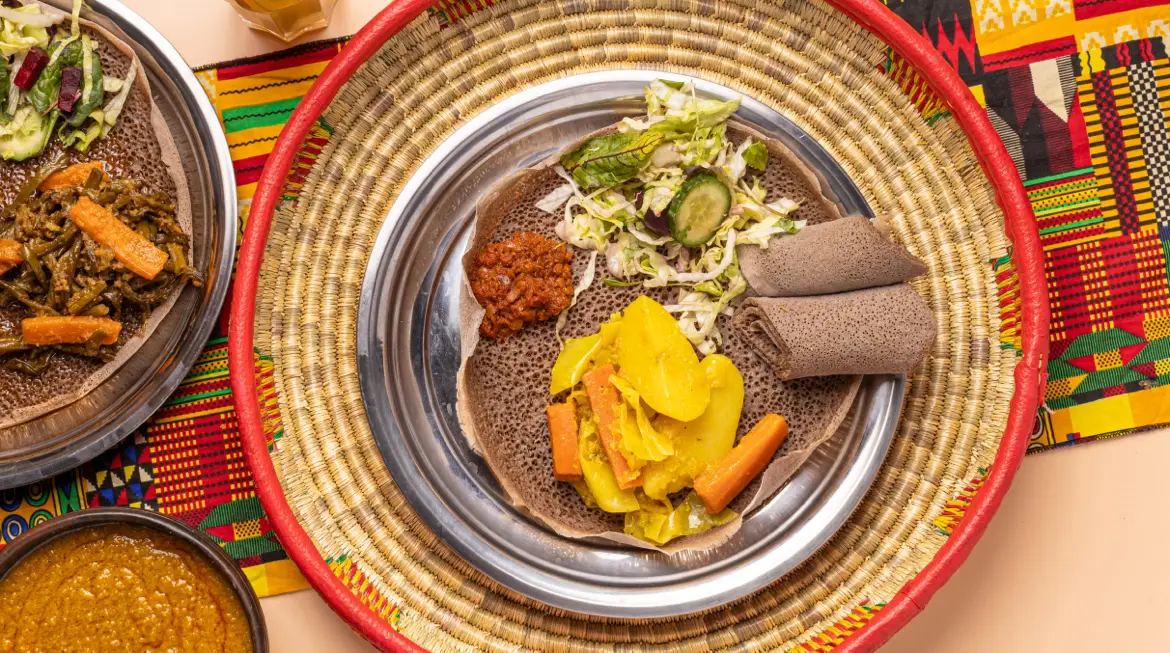 Injera-Teller von oben in einem geflochtenen Mesob: Salat, Kartoffel-Karotten-Gemüse, scharfe Soße und gerollte Injera